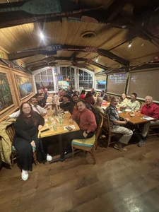 Large group of people around a long wooden table in a cozy rustic restaurant, smiling.