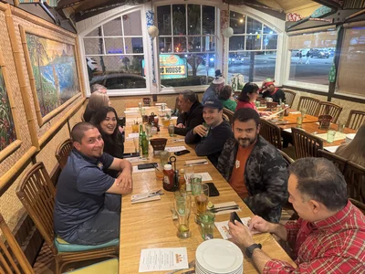 Group of people at a long wooden table in a tropical restaurant, with drinks, condiments and murals