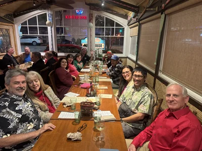 Long table packed with smiling diverse group in a cozy restaurant; menus and drinks visible.