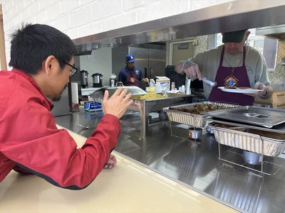 Man in red jacket watches server in maroon apron serving food from chafing trays at a cafeteria.