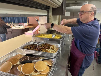 Cafeteria worker in navy shirt and maroon apron serves eggs; pancakes and sausages in front.