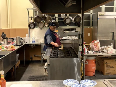 Chef in cap and apron grills at a commercial kitchen stove; hanging pots above stainless counters.