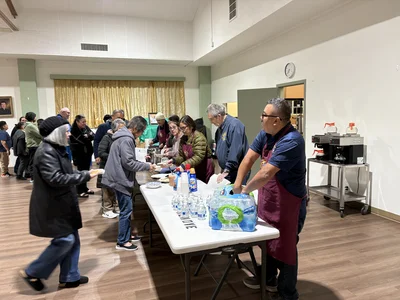 Community hall; volunteers in maroon aprons serve food to a line of people; coffee station on right.