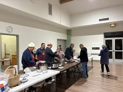 Volunteers in aprons serve meals from pots at a long table; clock, exit sign visible.