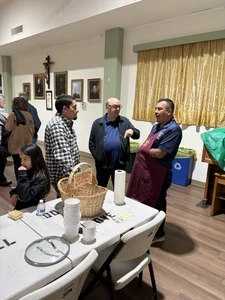 People chat at a table with a donation basket; portraits and a crucifix on the wall, beige curtains.