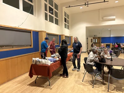 Community hall buffet: two servers at a red-covered table serving food; seniors dine at tables.